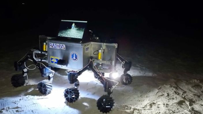 Robot with bright lights on a sandy lunar surface.