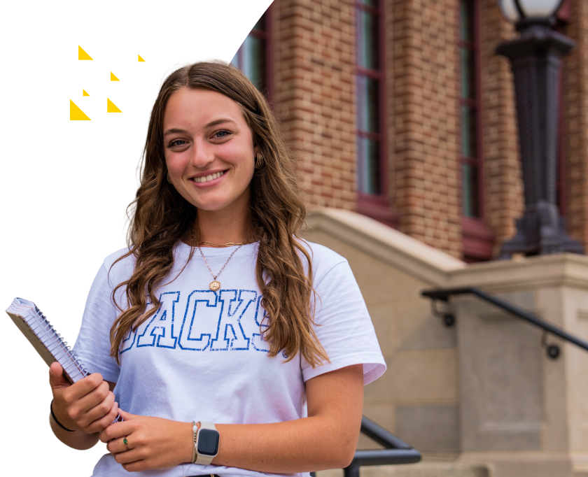 Smiling girl standing with notebook in front of campus building