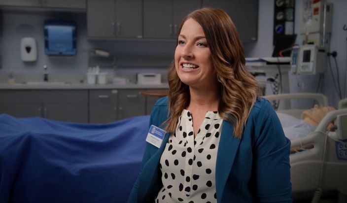 Female professor answering interview questions in a nursing simulation classroom with patient bed and equipment.